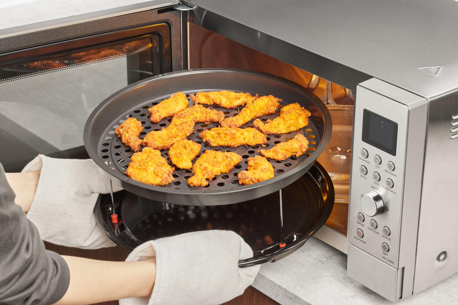 Panasonic NN-CD58RSEPG Panasonic NN-CD58RS kuchenka mikrofalowa Inverter Slim z grillem i funkcją Air Fry (1000W, termoobieg, 27l, kosz do smażenia beztłuszczowego), stalowa 26 Ręce w rękawicach wkładają do kuchenki Panasonic Slim Line Combi panierowane kawałki kurczaka w koszu Air Fry.